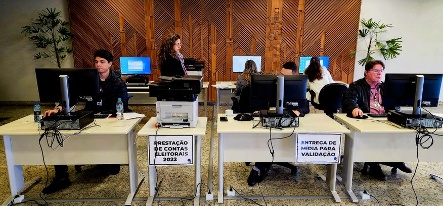 Fotografia de um ambiente com mesas de trabalho. Há pessoas sentadas diante de computadores e um...