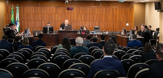 Foto em que aparecem os membros da Corte do TRE-PR, sentados atrás de mesas de madeira escura. M...