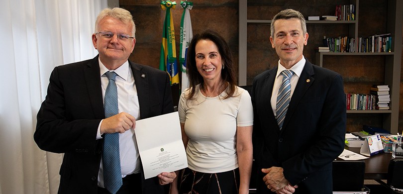 Fotografia de dois homens de terno e uma mulher de camisa branca posando sorrindo em uma sala co...