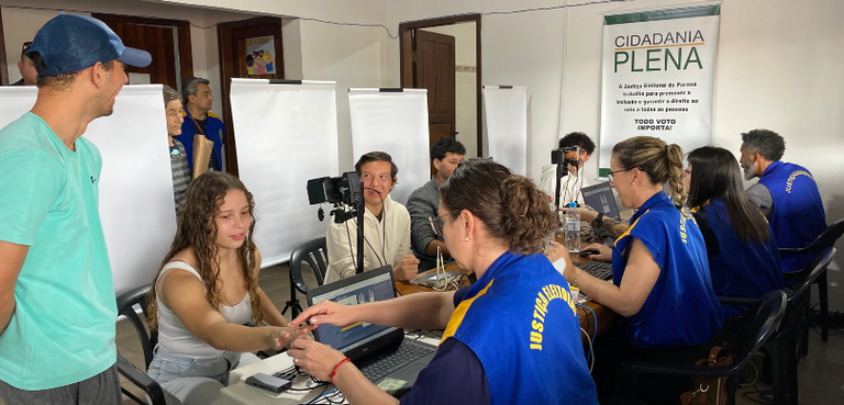 Programa “Cidadania Plena” atende 175 eleitores em Guaraqueçaba e região 2 Fotografia de quatro pessoas, que vestem o colete azul e amarelo da Justiça Eleitoral, realizand...