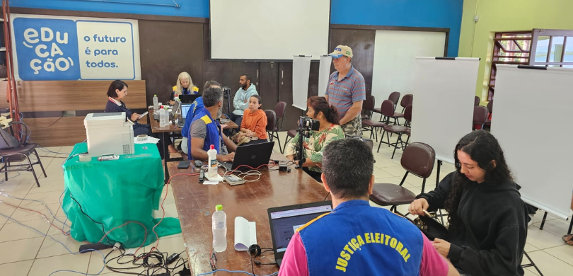Fotografia de ambiente interno onde ocorre uma ação de atendimento institucional. Em primeiro pl...