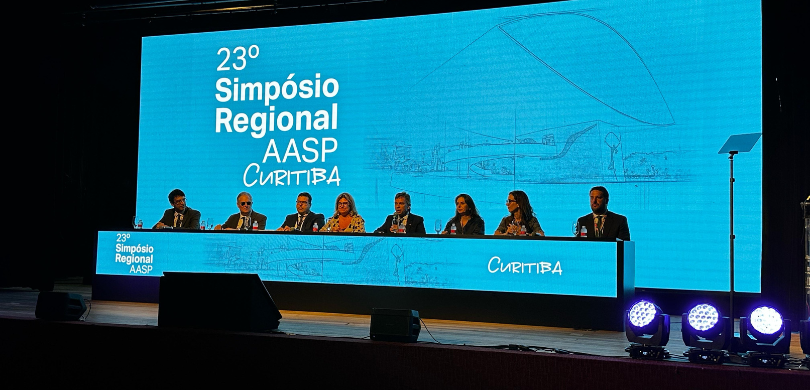 Fotografia da mesa de honra do evento, em um palco de auditório. É possível observar cinco homen...