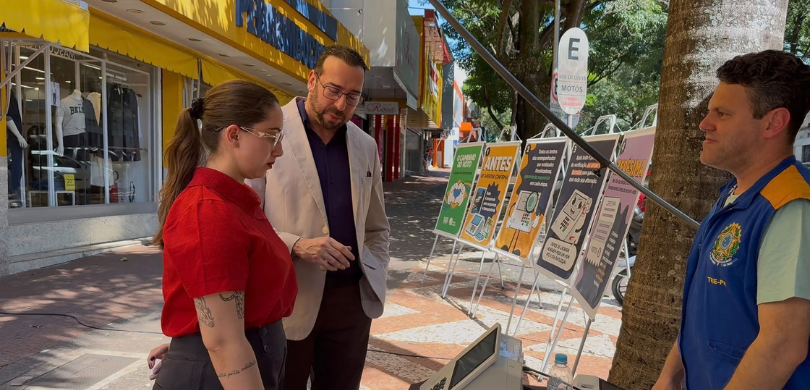 Fotografia em ambiente externo e calçado na qual se veem quatro pessoas em primeiro plano. À esq...
