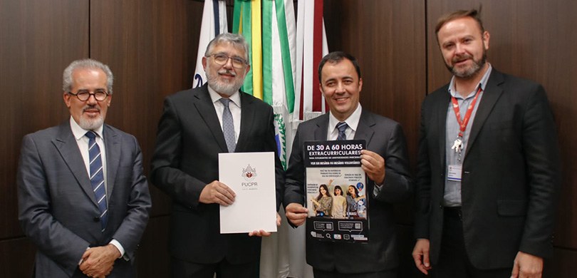 Fotografia de quatro homens, um ao lado do outro. No centro, dois deles mostram os papéis que es...