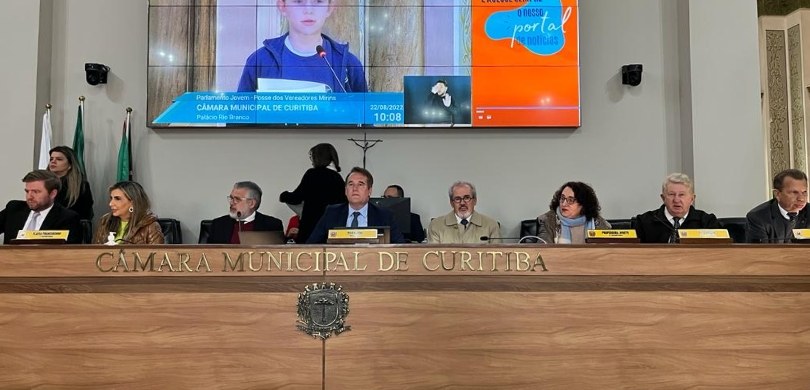 Fotografia da mesa principal da Câmara Municipal de Curitiba. Oito pessoas estão sentadas, sendo...