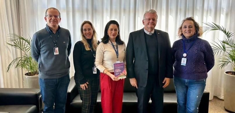 Fotografia em que aparecem três mulheres e dois homens posando em uma sala. Entre essas pessoas ...