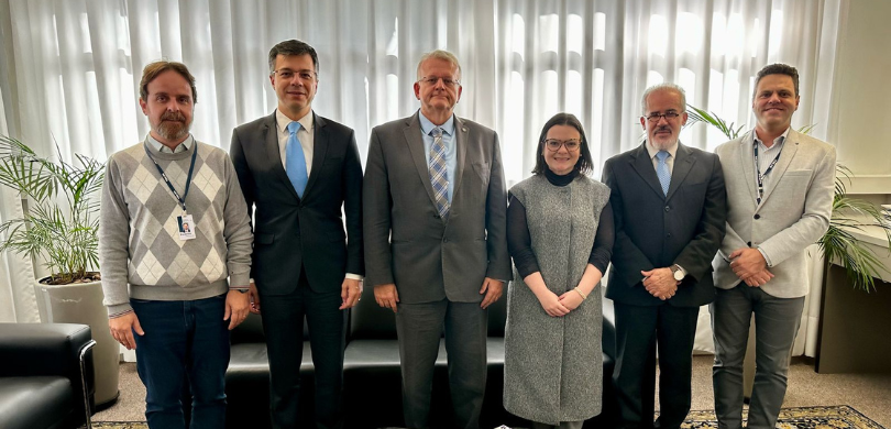 Fotografia de cinco homens e uma mulher posando sorrindo em uma sala com paredes brancas e piso ...