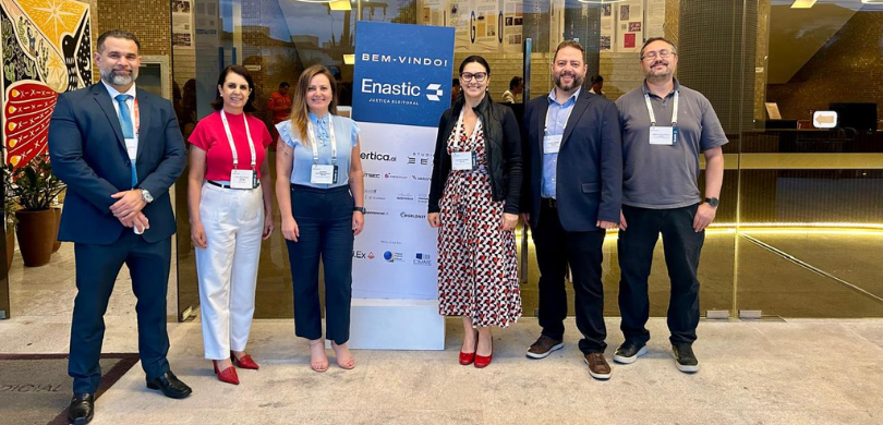 Fotografia de três mulheres e três homens posando sorrindo ao redor de um banner branco e azul e...