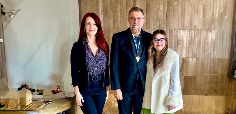 Em uma sala com paredes brancas e bege, duas mulheres e um homem posam para a foto. O homem está...