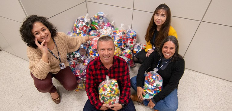 Fotografia de quatro pessoas, três mulheres e um homem, sentados no chão ao redor de vários paco...