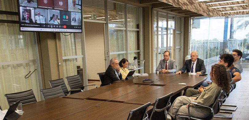 Fotografia de uma reunião híbrida em sala institucional. Ao redor de uma longa mesa de madeira e...