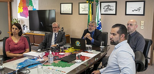 Reunião com zonas eleitorais do Paraná alinha ações para as Eleições 2022 Fotografia de pessoas sentadas ao redor de uma mesa de trabalho. Ao fundo, há a bandeira do Bras...