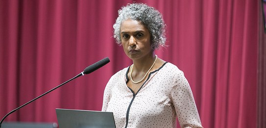 A fotografia mostra uma televisão e atrás um painel e uma cortina da cor vermelha. A tevê mostra...