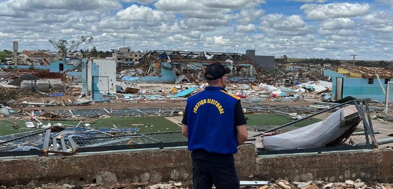 Fotografia em que se observa, de costas, um homem vestindo um colete azul e amarelo da Justiça E...