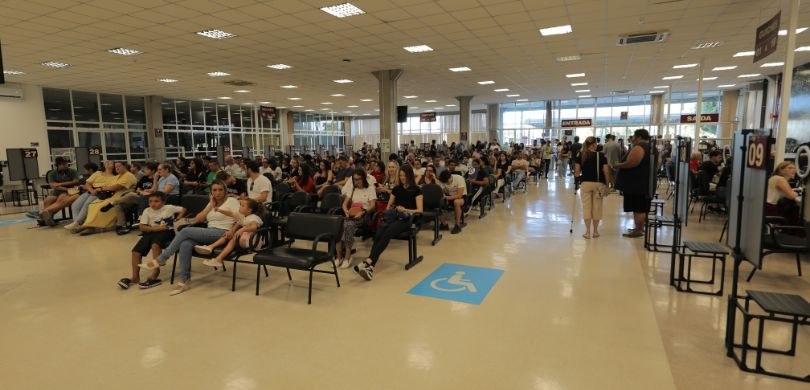 Fotografia de uma ampla sala de espera interna, com diversas pessoas, entre homens, mulheres e c...