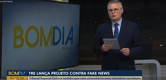 Reprodução de telejornal. Há um apresentador de terno e gravata à direita ao lado de uma tela on...