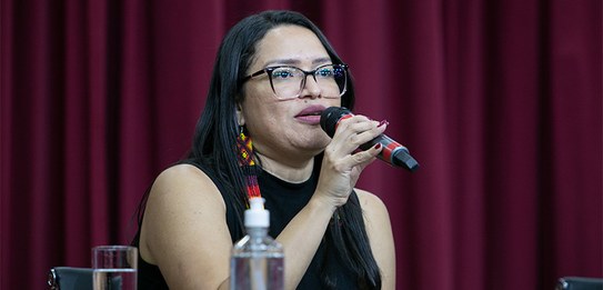 A fotografia mostra uma televisão e atrás um painel e uma cortina da cor vermelha. A tevê mostra...