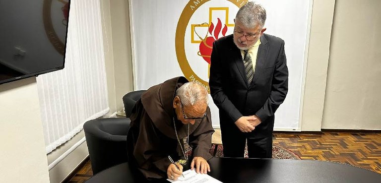 No centro da imagem, um homem vestindo um hábito religioso está agachado assinando um papel. Do ...