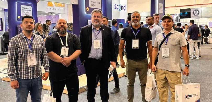Fotografia de cinco homens posando sorrindo em frente aos stands da “Feira - ISC Security Evento...