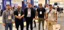 Fotografia de cinco homens posando sorrindo em frente aos stands da “Feira - ISC Security Evento...