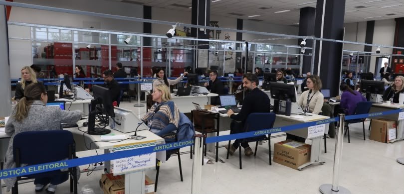 Fotografia que retrata o interior de um amplo salão de atendimento com diversas pessoas, entre h...
