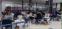 Fotografia que retrata o interior de um amplo salão de atendimento com diversas pessoas, entre h...