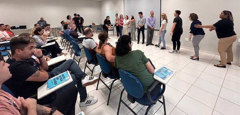 Em uma sala clara bastante ampla, várias pessoas enfileiradas são observadas por outras pessoas ...