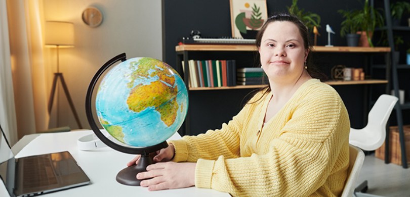 Mulher vestindo uma blusa amarela segura um globo terrestre enquanto está sentada em uma escriva...