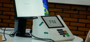 Fotografia na qual se vê uma urna eletrônica de cor clara e um monitor de computador dispostos s...
