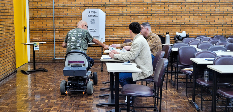 Fotografia na qual se observa uma Seção Eleitoral organizada em um ambiente interno. Ao centro, ...