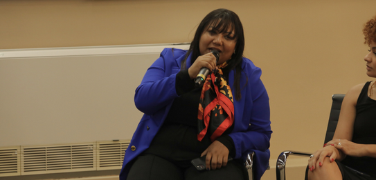 Fotografia de uma mulher negra de cabelos crespos curtos sorrindo segurando um microfone. Ela ve...