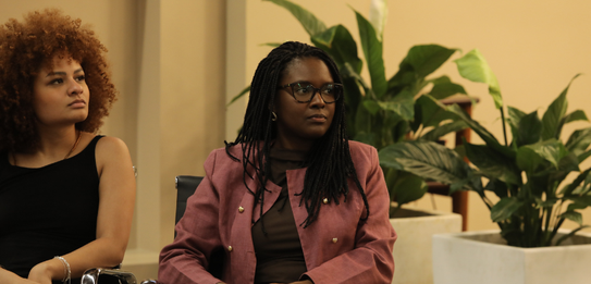 Fotografia de uma mulher negra de cabelos crespos curtos sorrindo segurando um microfone. Ela ve...