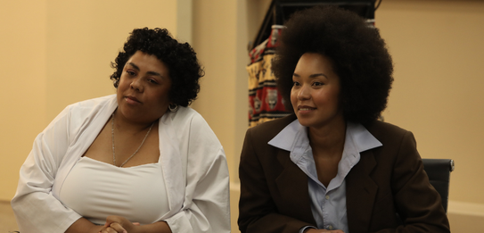 Fotografia de uma mulher negra de cabelos crespos curtos sorrindo segurando um microfone. Ela ve...