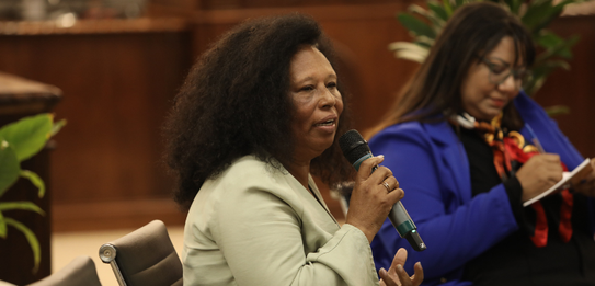 Fotografia de uma mulher negra de cabelos crespos curtos sorrindo segurando um microfone. Ela ve...