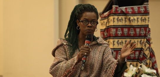 Fotografia de uma mulher negra de cabelos crespos curtos sorrindo segurando um microfone. Ela ve...