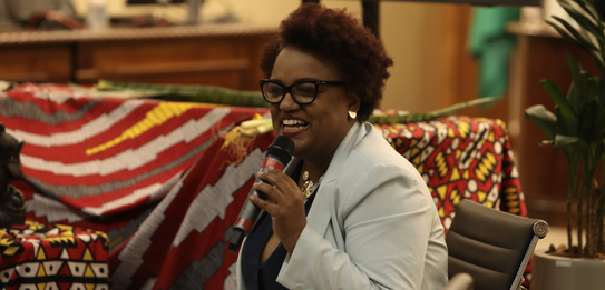 Fotografia de uma mulher negra de cabelos crespos curtos sorrindo segurando um microfone. Ela ve...