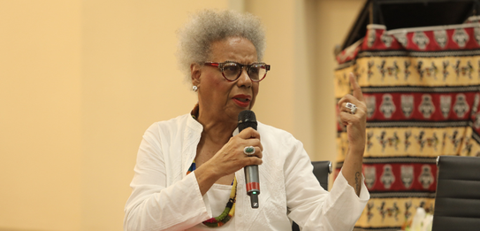Fotografia de uma mulher negra de cabelos crespos curtos sorrindo segurando um microfone. Ela ve...