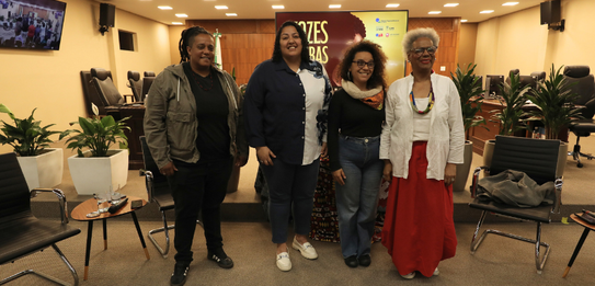 Fotografia de uma mulher negra de cabelos crespos curtos sorrindo segurando um microfone. Ela ve...