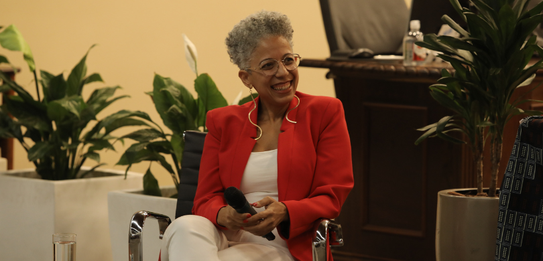 Fotografia de uma mulher negra de cabelos crespos curtos sorrindo segurando um microfone. Ela ve...