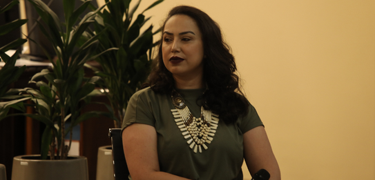 Fotografia de uma mulher negra de cabelos crespos curtos sorrindo segurando um microfone. Ela ve...