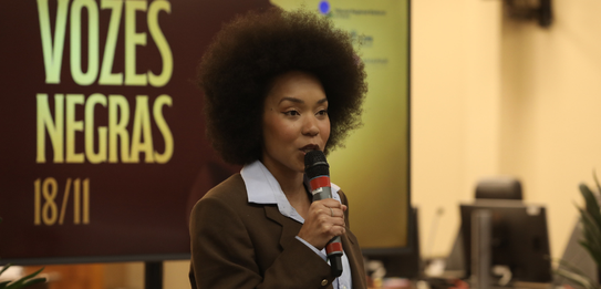 Fotografia de uma mulher negra de cabelos crespos curtos sorrindo segurando um microfone. Ela ve...