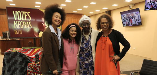 Fotografia de uma mulher negra de cabelos crespos curtos sorrindo segurando um microfone. Ela ve...