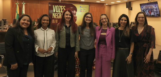Fotografia de uma mulher negra de cabelos crespos curtos sorrindo segurando um microfone. Ela ve...