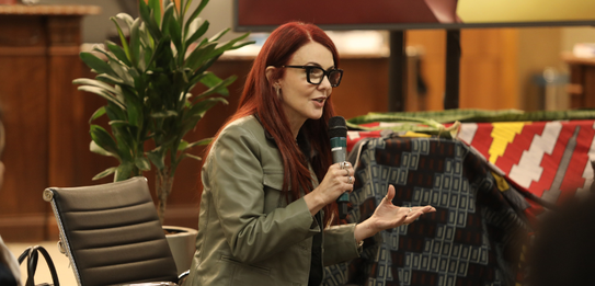 Fotografia de uma mulher negra de cabelos crespos curtos sorrindo segurando um microfone. Ela ve...
