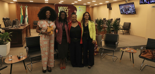 Fotografia de uma mulher negra de cabelos crespos curtos sorrindo segurando um microfone. Ela ve...