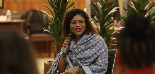 Fotografia de uma mulher negra de cabelos crespos curtos sorrindo segurando um microfone. Ela ve...