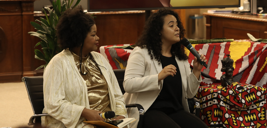 Fotografia de uma mulher negra de cabelos crespos curtos sorrindo segurando um microfone. Ela ve...