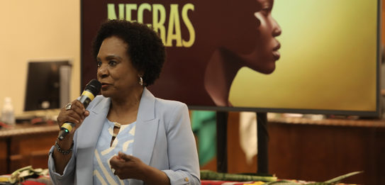 Fotografia de uma mulher negra de cabelos crespos curtos sorrindo segurando um microfone. Ela ve...