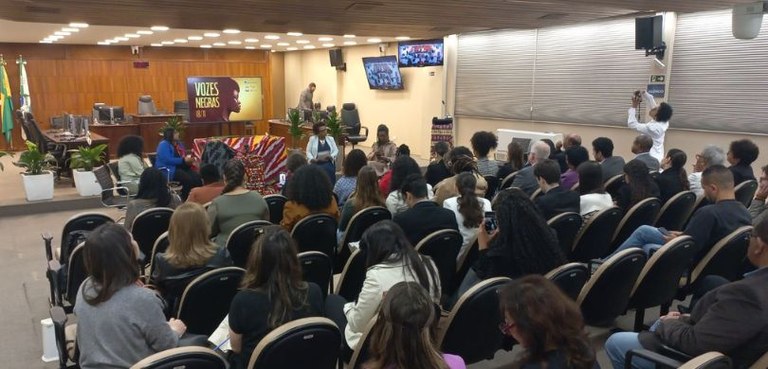 Fotografia de diversas pessoas, entre homens e mulheres, sentados em cadeiras pretas com estofad...