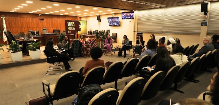 Fotografia de sete mulheres sentadas em cadeiras pretas, organizadas em meio círculo, durante a ...